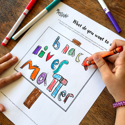 Closeup of a kid, hands only visible, colouring the "Design Your Own T-shirt" activity sheet with crayons and markers